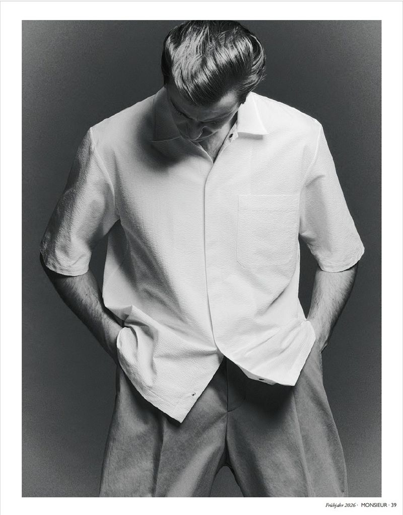 Model Ben Allen wearing a white short-sleeve shirt with hands in pockets against a grey studio backdrop for Monsieur Magazin Germany.