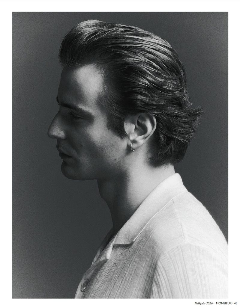 Profile portrait of a male model in a white shirt with slicked back hair and a small hoop earring against a grey studio backdrop.