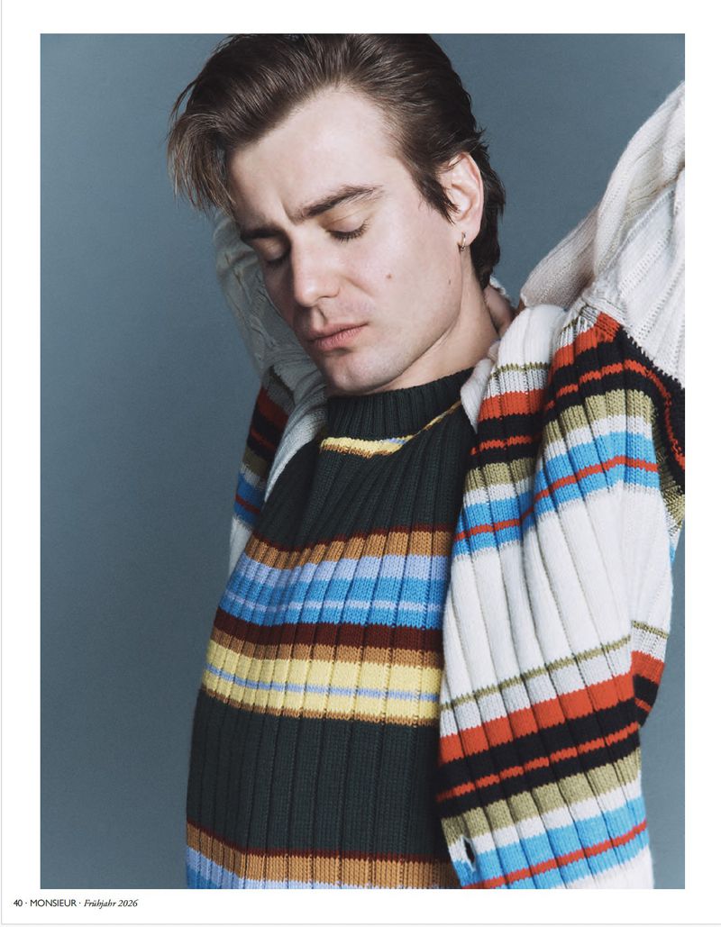 Man wearing a striped knit sweater stretching arms overhead against a grey background