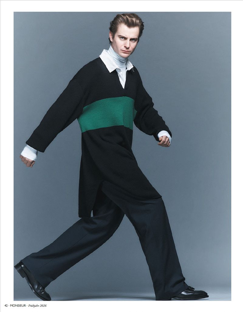 Model wearing a black tunic with a green stripe over dark trousers against a grey backdrop for Monsieur Magazin.