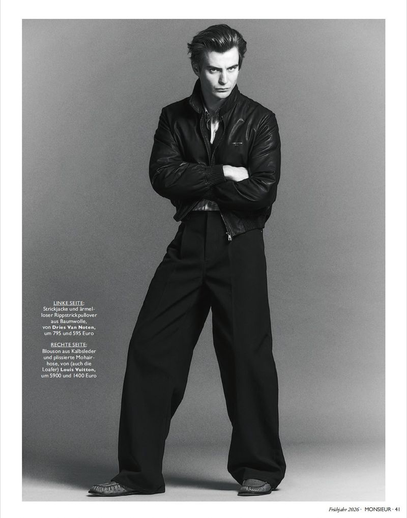 Full-length black and white editorial portrait of a male model with arms crossed wearing a leather jacket and wide trousers.