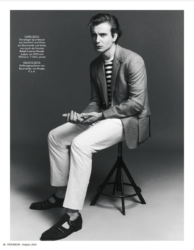 Seated male model wearing grey blazer, striped t-shirt and white trousers on a stool in a studio setting