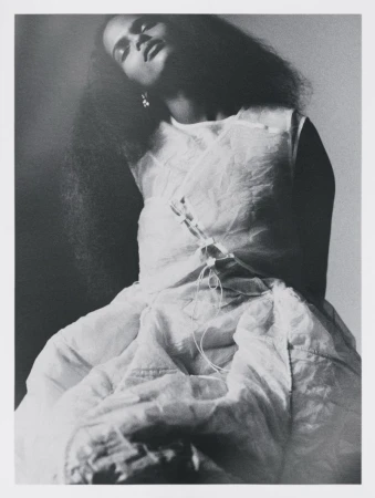 Black and white portrait of a model with voluminous dark hair wearing a translucent layered dress against a plain studio background.