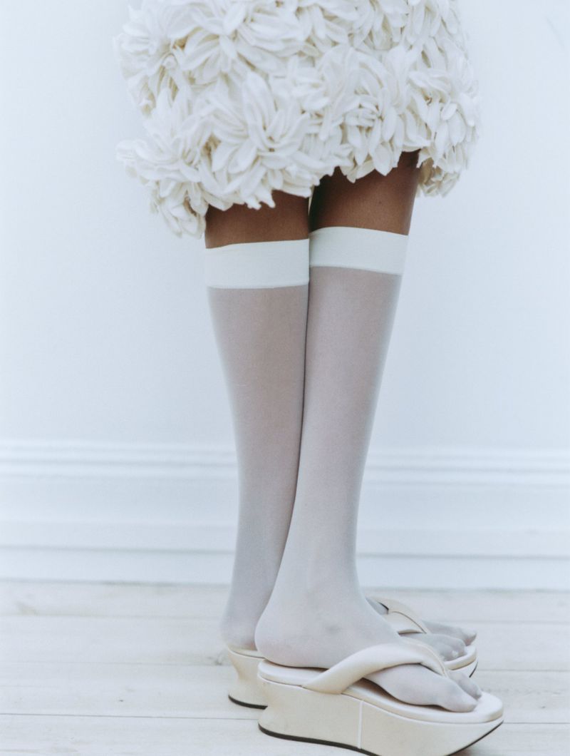 Close-up of legs wearing white knee-high sheer socks and platform sandals under a textured white miniskirt against a plain background