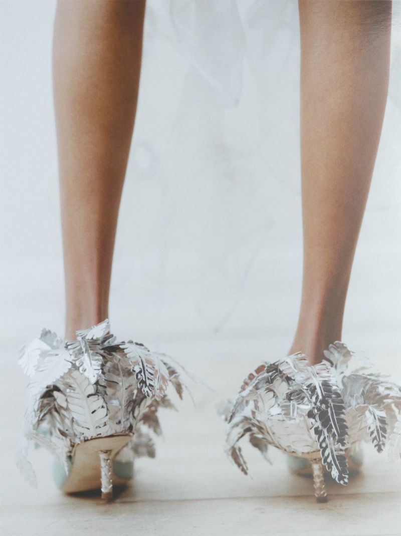 Close-up of bare legs wearing ornate silver leaf-embellished high heel shoes against a white studio background.