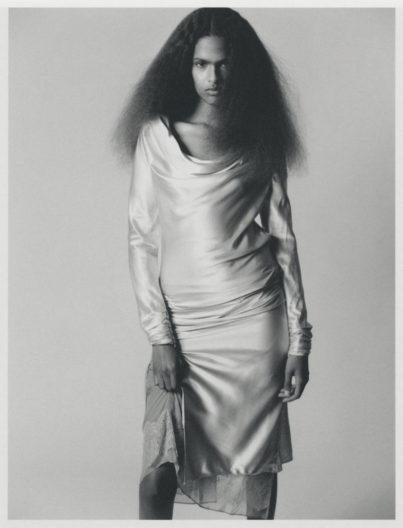 Model with voluminous dark hair wearing a long draped silk dress against a neutral grey studio backdrop