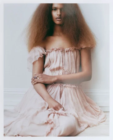 Model in a pale pink ruffled dress with voluminous hair seated against a plain white background for Manifesto Magazine.
