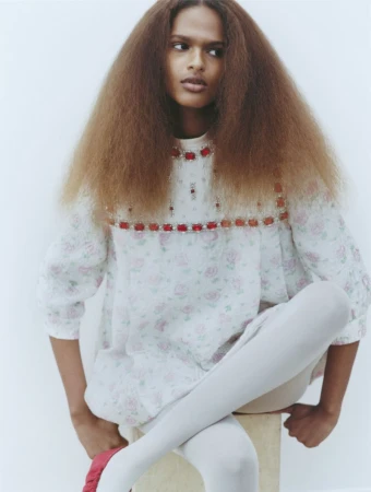 Model with voluminous brown hair wearing a floral embellished top and white tights sitting on a light backdrop