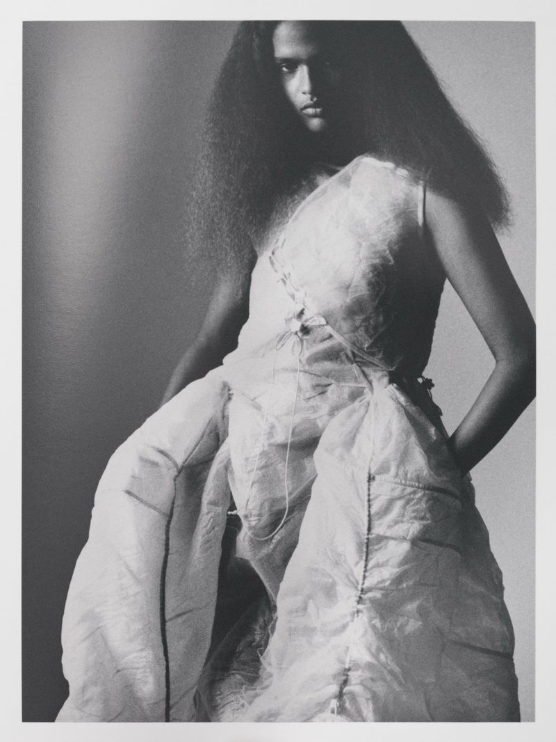 Fashion shot of a female model in a puffy white dress with dark, curly hair against a plain gray background in a studio setting.