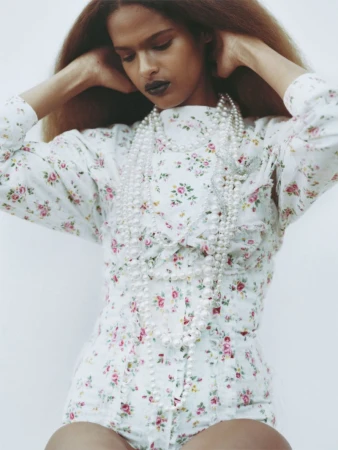 Model in white floral romper wearing layered pearl necklaces against a seamless pale background for Manifesto Magazine.