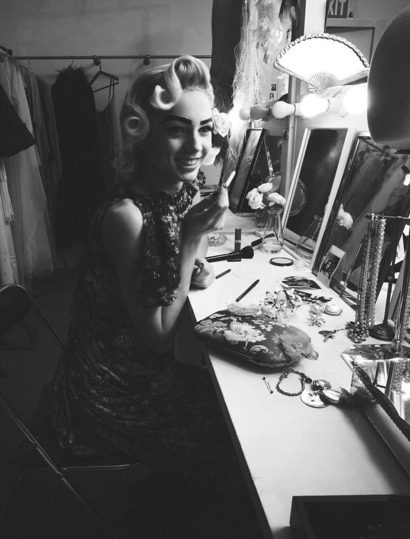 Model with vintage hairstyle seated at a brightly lit dressing room vanity table applying makeup