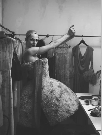 Model in a voluminous textured gown posing in a backstage wardrobe room filled with hanging garments