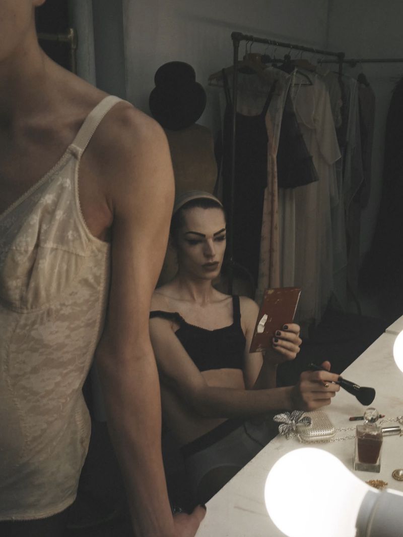 Model seated in front of a mirror holding a makeup brush in a dressing room setting for System Magazine.