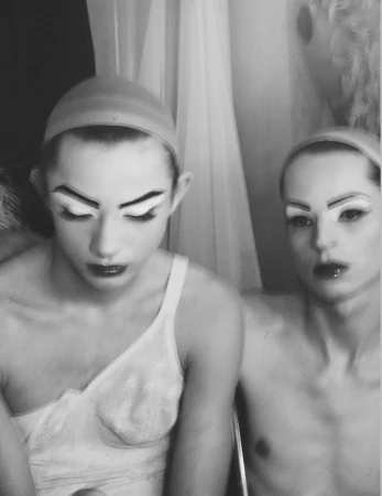 Two male models in white undergarments and bald caps with theatrical stage makeup in a black and white portrait.