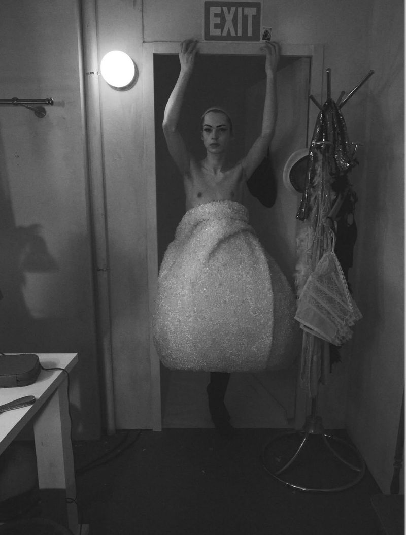 Model standing in a doorway wearing a sculptural textured garment with arms raised holding the frame, shot by David Sims for System Magazine