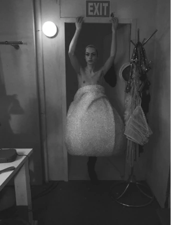 Model standing in a doorway wearing a sculptural textured garment with arms raised holding the frame, shot by David Sims for System Magazine