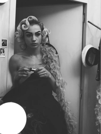 Model with vintage styled blonde hair and feather boa holding a lipstick in a backstage dressing room setting
