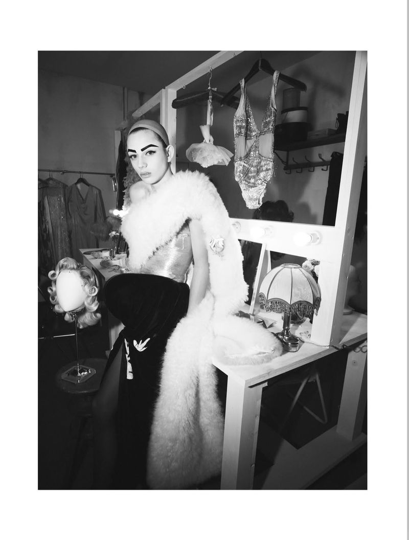 Model wearing a feathered wrap and dark skirt standing in a dressing room backstage for System Magazine