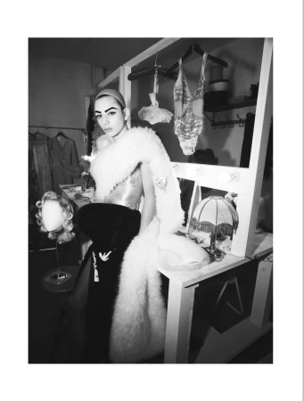 Model wearing a feathered wrap and dark skirt standing in a dressing room backstage for System Magazine