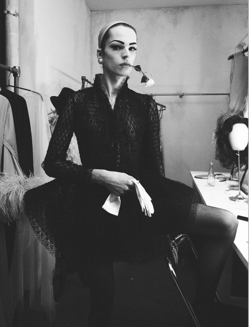 Model in dark lace dress holding a white flower in teeth and white gloves in hand inside a backstage dressing room