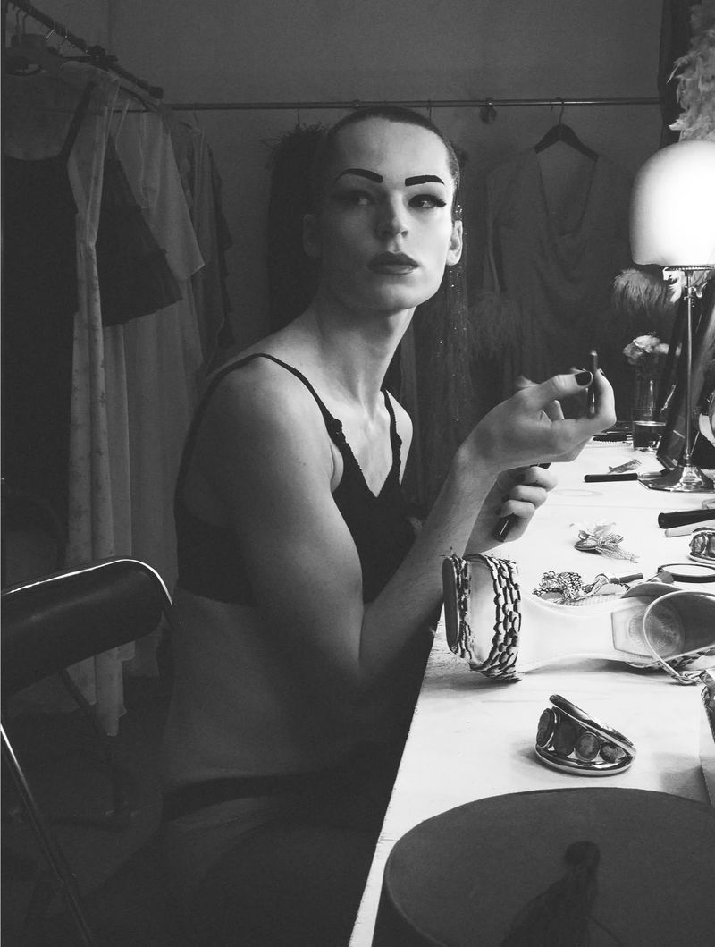 Model seated at a dressing table holding a cosmetic pencil in a backstage setting captured in black and white.