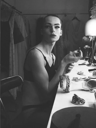 Model seated at a dressing table holding a cosmetic pencil in a backstage setting captured in black and white.