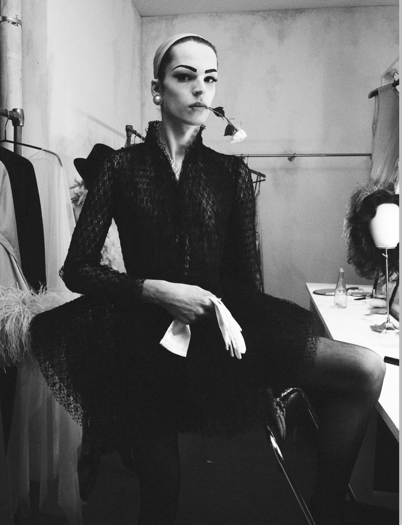 Model in black lace dress holding a white rose and gloves in dressing room setting