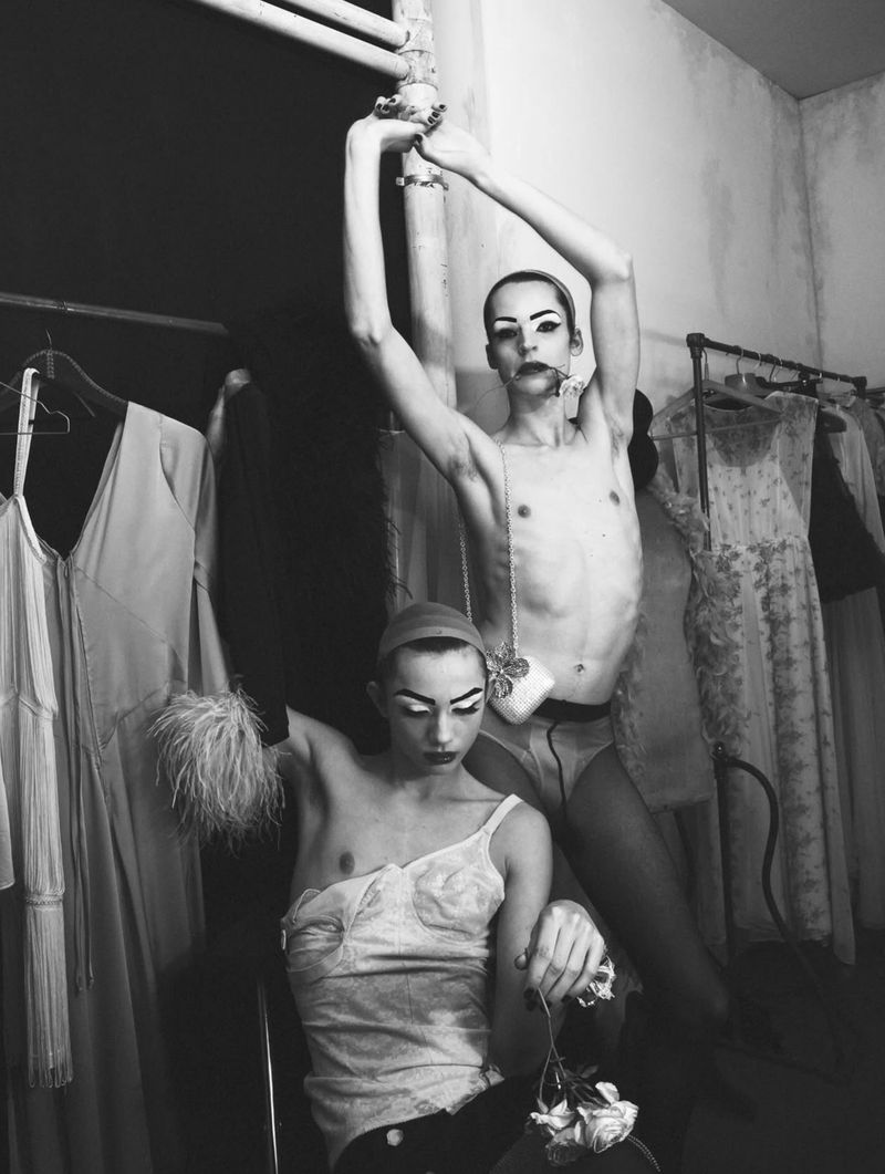 Black and white editorial photograph of two male models posing backstage amongst clothing racks in a dressing room for System Magazine.
