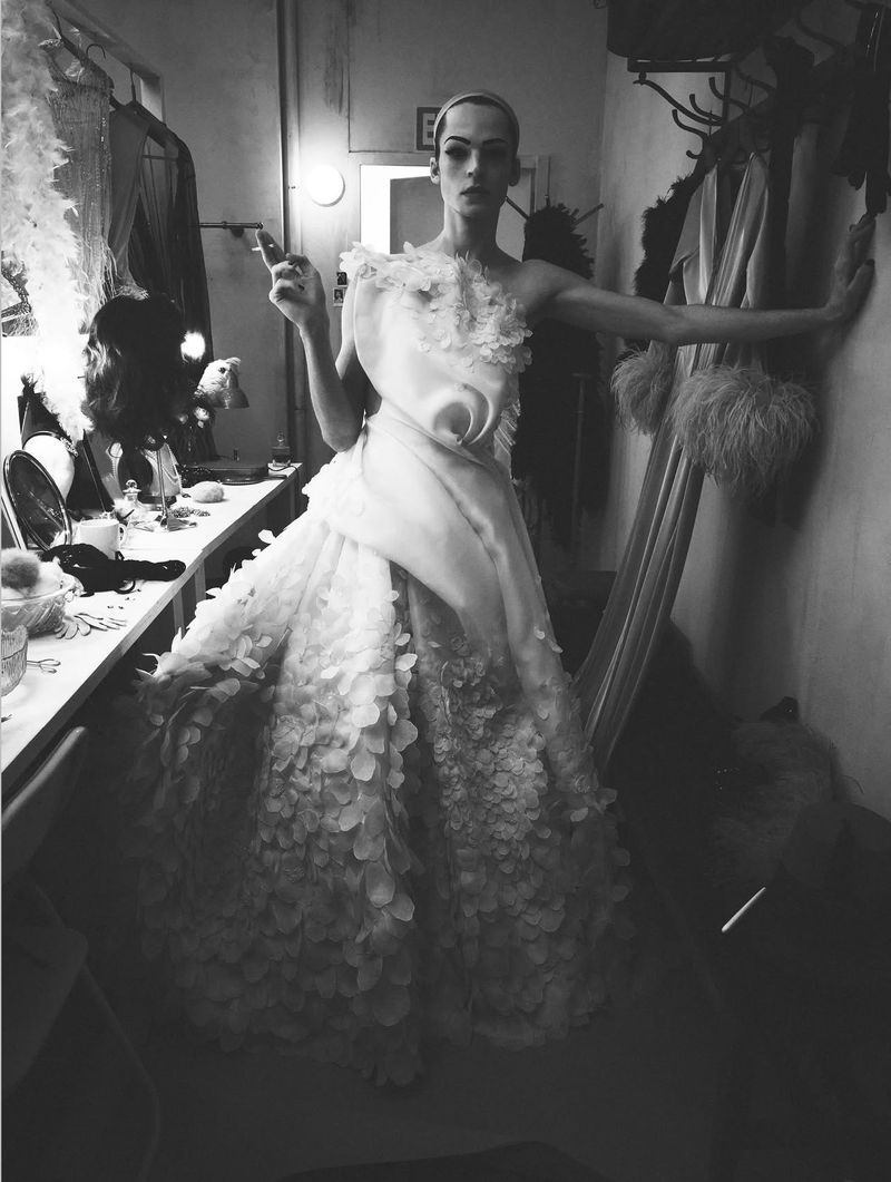 Model in a sculptural white gown posing in a backstage dressing room holding a cigarette for System Magazine.