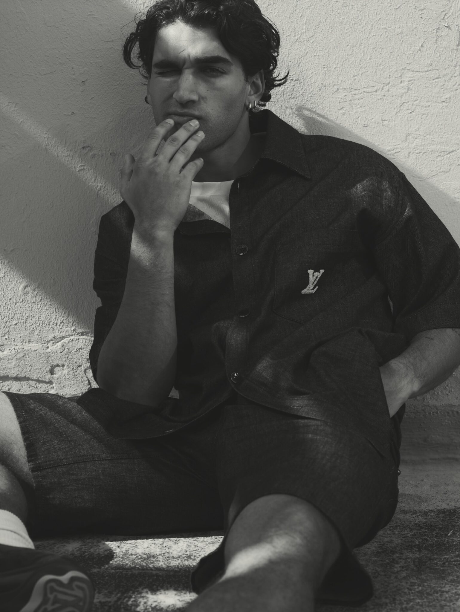 Monochrome portrait of a young adult man with dark hair, wearing an open shirt, sitting in an outdoor setting, with hand to face.