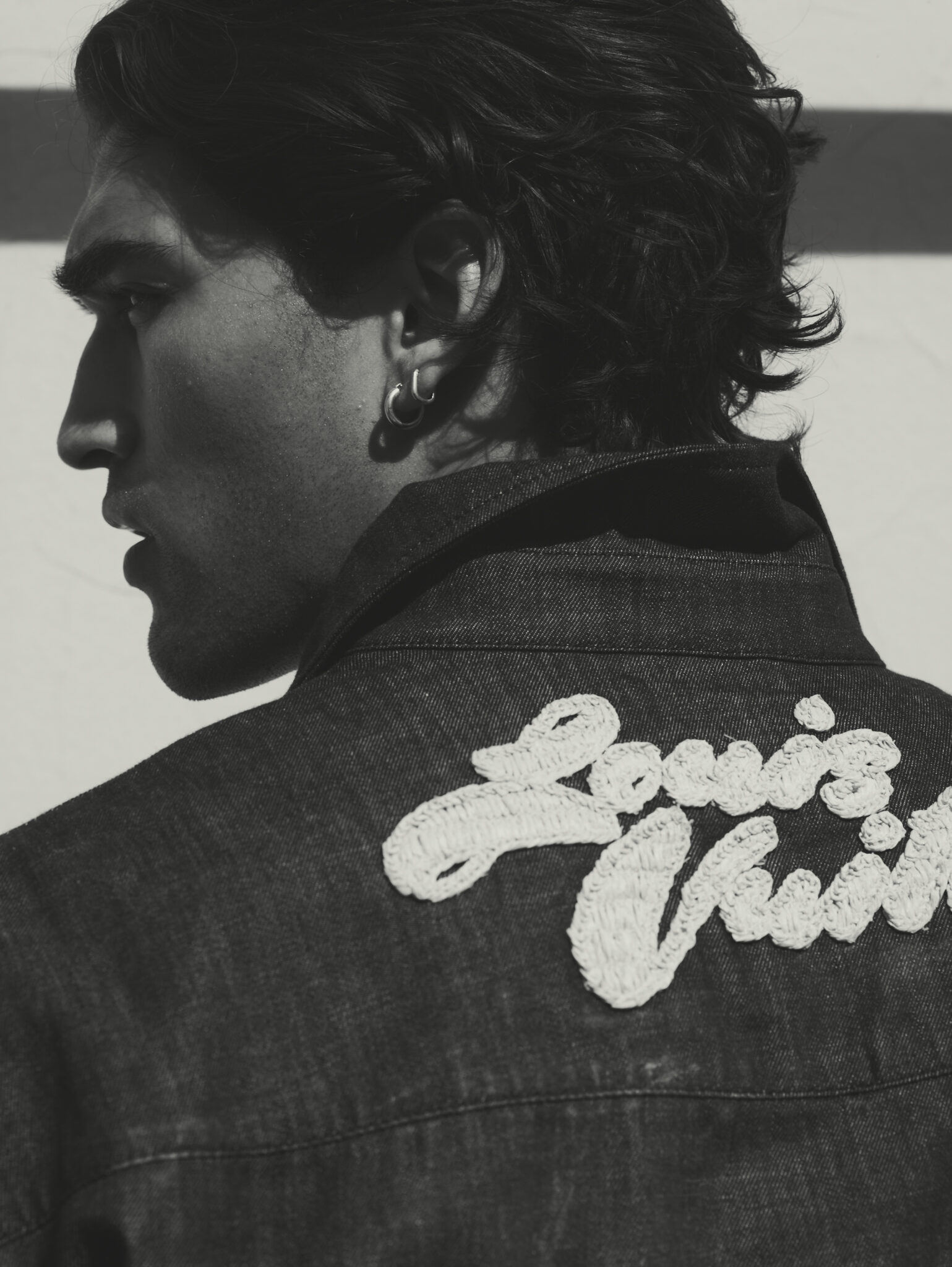 Monochrome portrait of a stylish adult male with dark hair in a denim jacket featuring an embroidered 'Louis Vuitton' logo on the back.