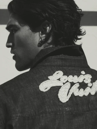 Monochrome portrait of a stylish adult male with dark hair in a denim jacket featuring an embroidered 'Louis Vuitton' logo on the back.