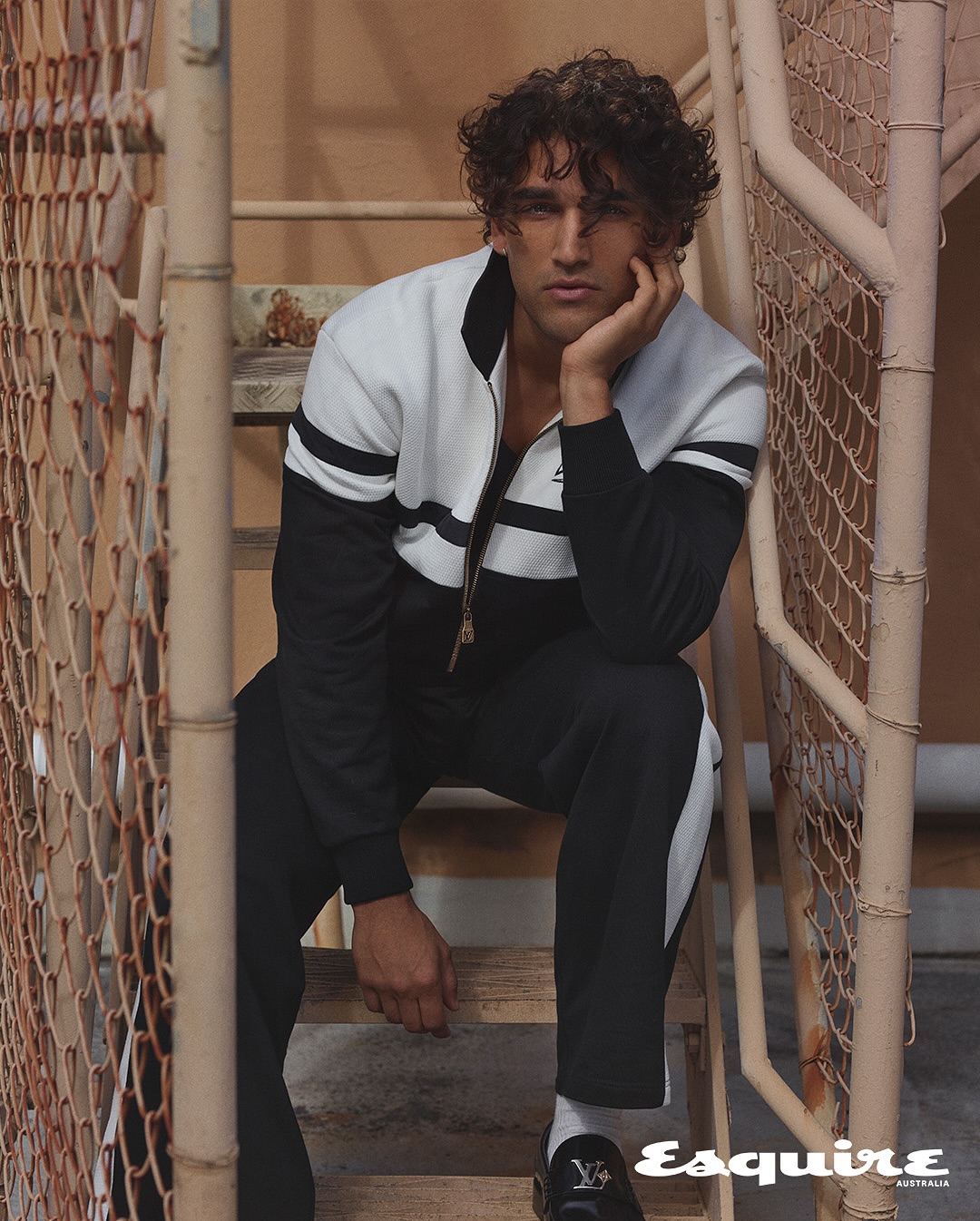 A male model sits on metal stairs in a black and white track suit, accessorized with black loafers and white socks, for a fashion editorial.