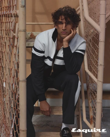 A male model sits on metal stairs in a black and white track suit, accessorized with black loafers and white socks, for a fashion editorial.