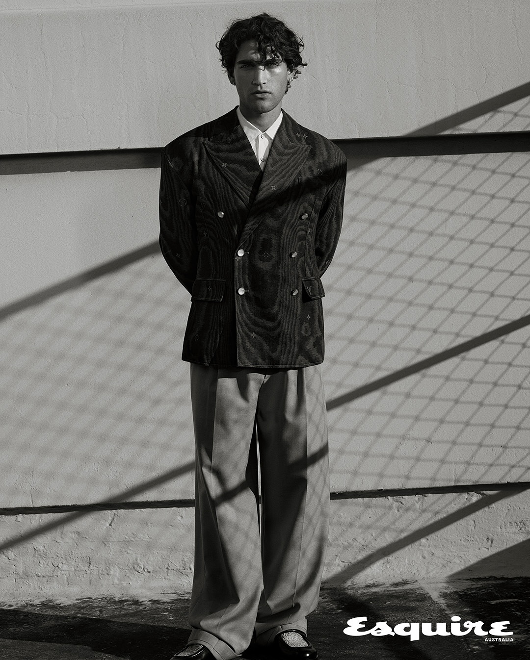 A young adult man poses in a double-breasted jacket and wide-leg trousers against a wall with geometric shadows in a black-and-white fashion shot.
