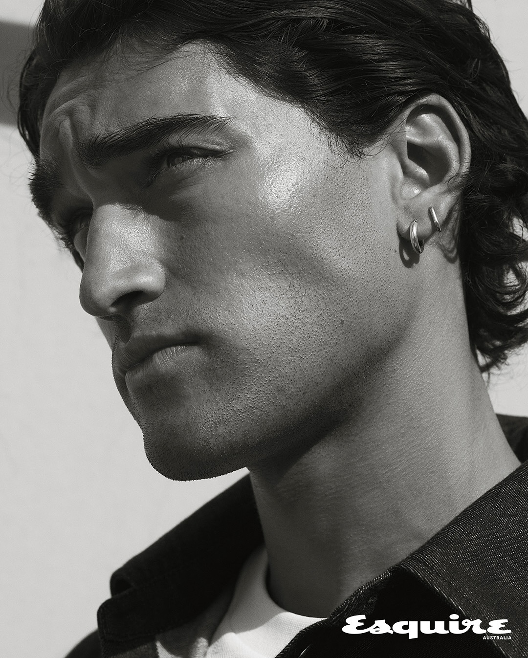 Monochrome portrait of a handsome adult male model with dark hair, hoop earrings, and wearing a dark denim jacket against a plain backdrop.