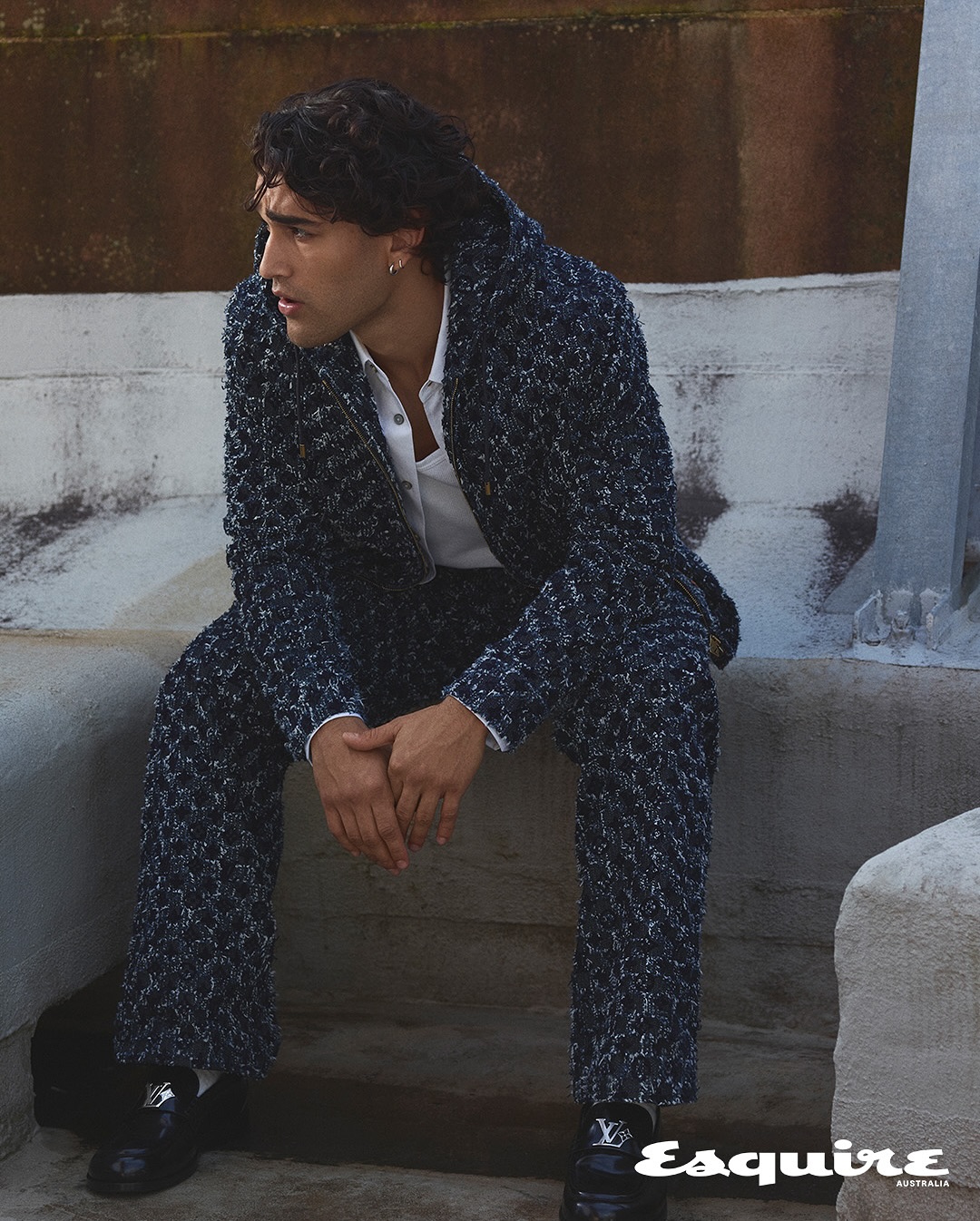 Man in textured suit sits on steps outdoors, looking away. Stylish formal wear fashion editorial with natural light and Esquire logo.