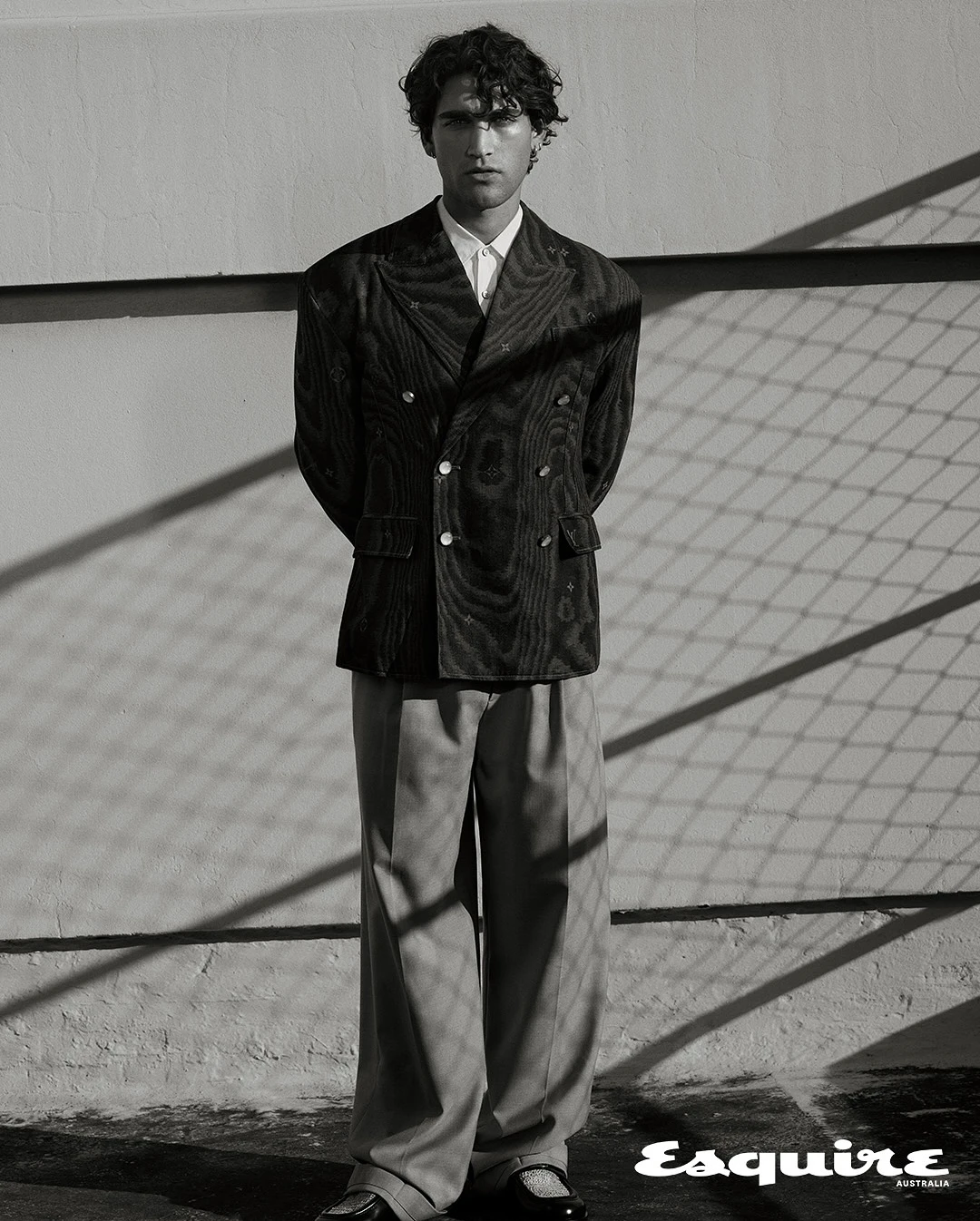 Man wearing a textured double-breasted blazer and trousers standing against a wall with chain-link shadows