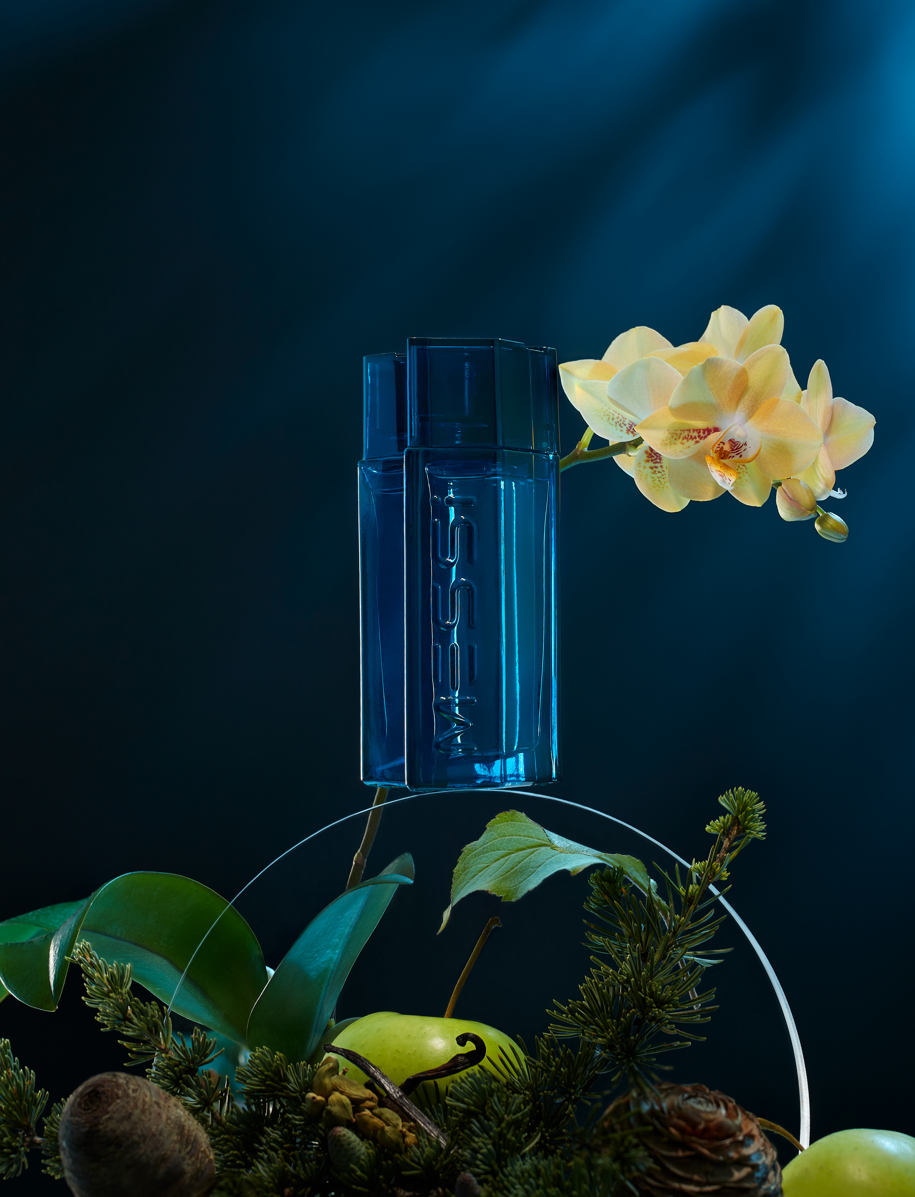 Blue glass perfume bottle on a clear stand with orchid flower, apple, and greenery against a dark teal backdrop with light.
