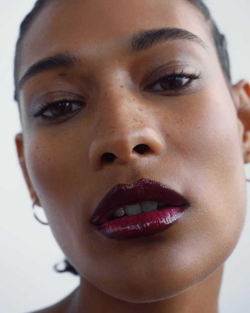 Close-up shot of a young adult woman with dark hair and skin wearing a shiny, dark red lip gloss in a brightly lit, neutral setting.