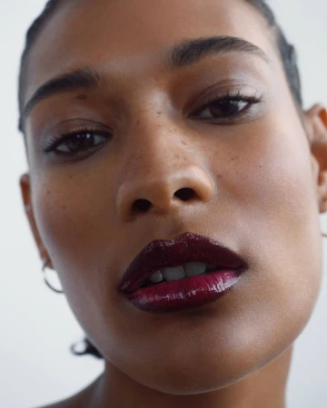 Close-up shot of a young adult woman with dark hair and skin wearing a shiny, dark red lip gloss in a brightly lit, neutral setting.