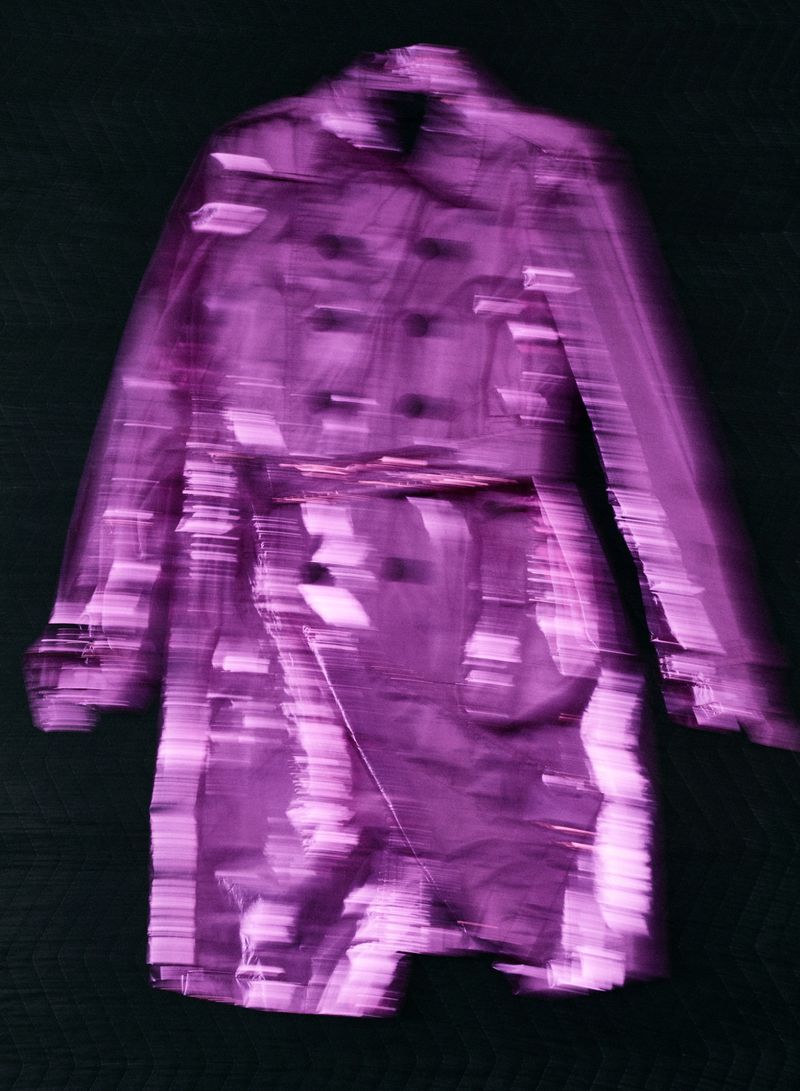 Blurry long purple trench coat displayed centrally against a dark background in an editorial fashion photograph.
