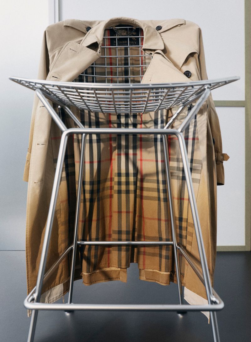 Tan trench coat hung over the back of a modern wire mesh chair in a studio setting.