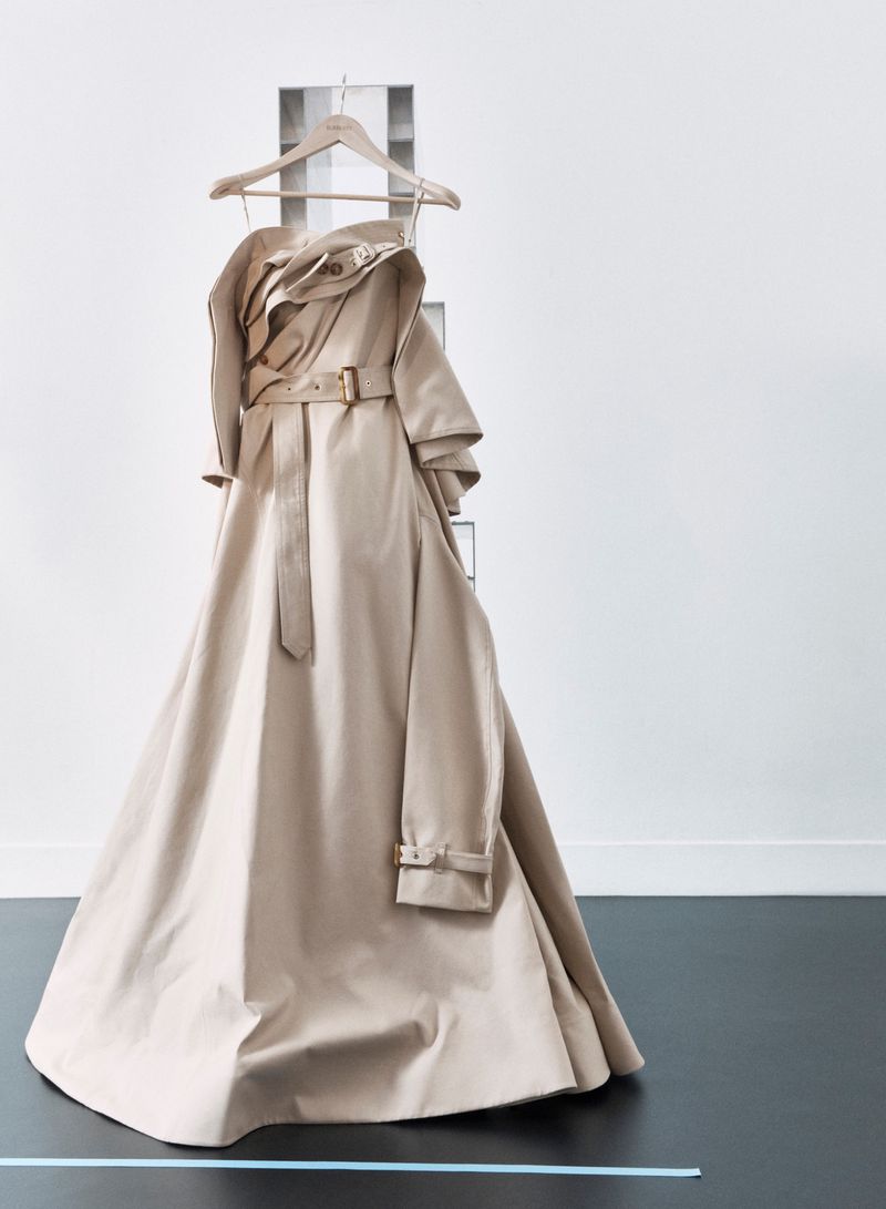 Beige trench coat on hanger against white wall with gray ladder-like display. Fashionable outerwear in minimalist studio setting.
