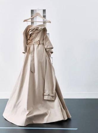 Beige trench coat on hanger against white wall with gray ladder-like display. Fashionable outerwear in minimalist studio setting.