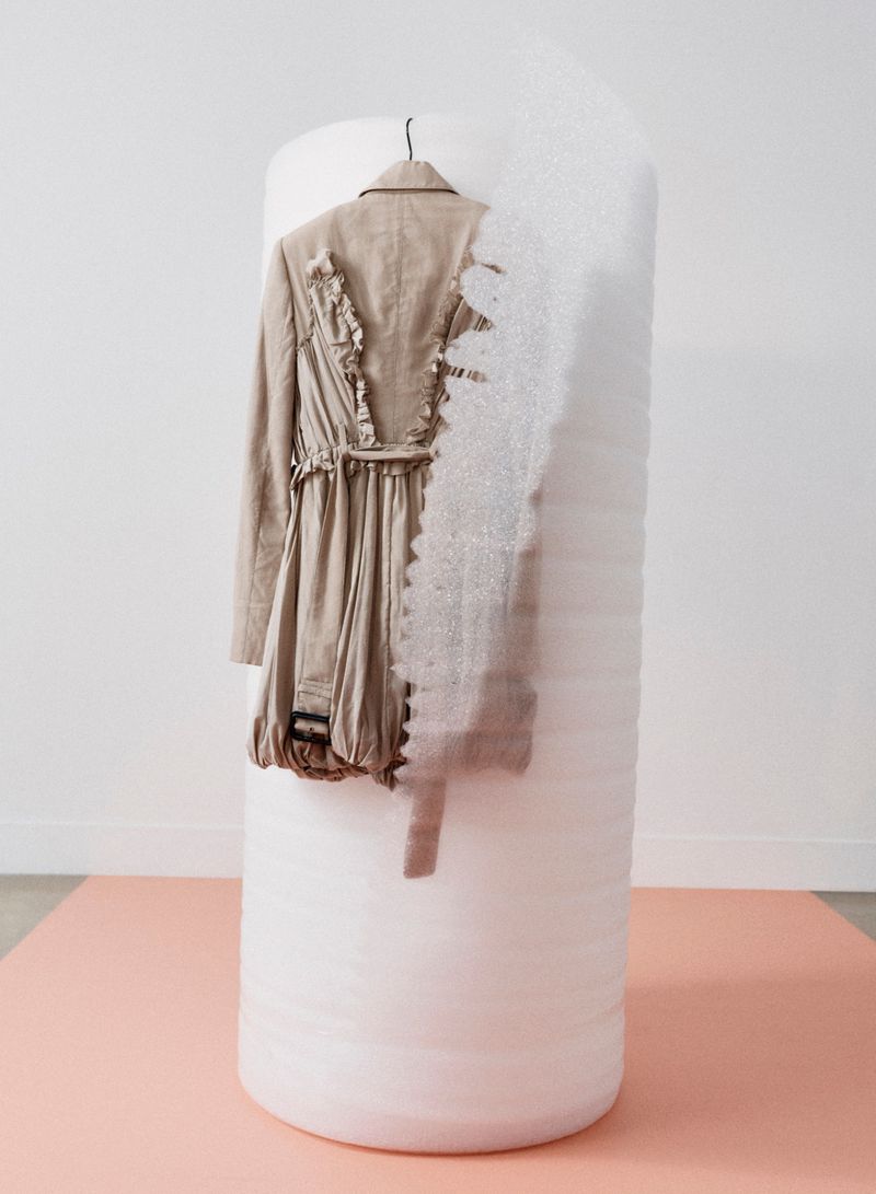 Beige trench coat hanging on a vertical white cylindrical architectural form against a plain white wall and peach floor.