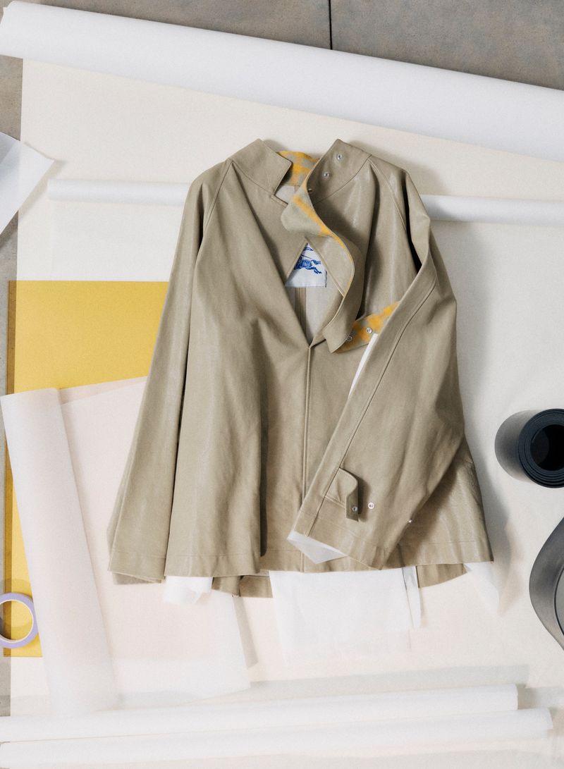 A beige trench coat jacket displayed flat against a background of rolled white paper and colored sheets.
