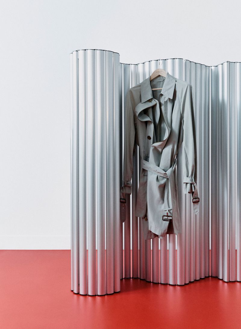A grey trench coat hanging on a wooden hanger against a corrugated metallic screen and a red floor.