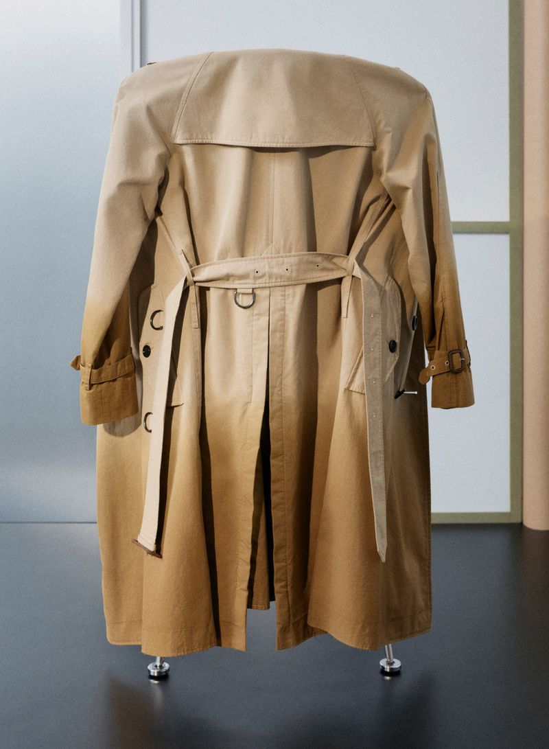 Tan belted Burberry trench coat displayed on a sculptural form against an interior architectural background