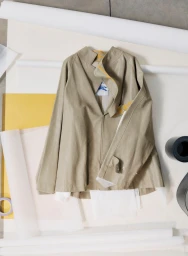 A beige trench coat jacket displayed flat against a background of rolled white paper and colored sheets.